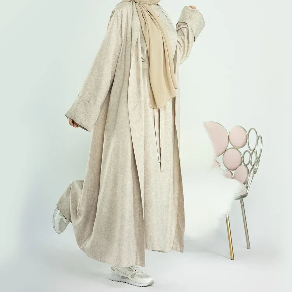 Woman wearing beige abaya with beige hijab and white sneakers, standing near modern chair with pink cushions