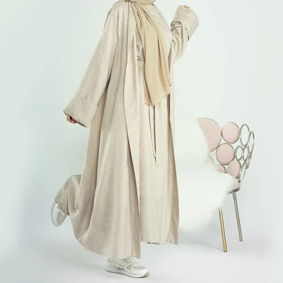 Woman wearing beige abaya with beige hijab and white sneakers, standing near modern chair with pink cushions